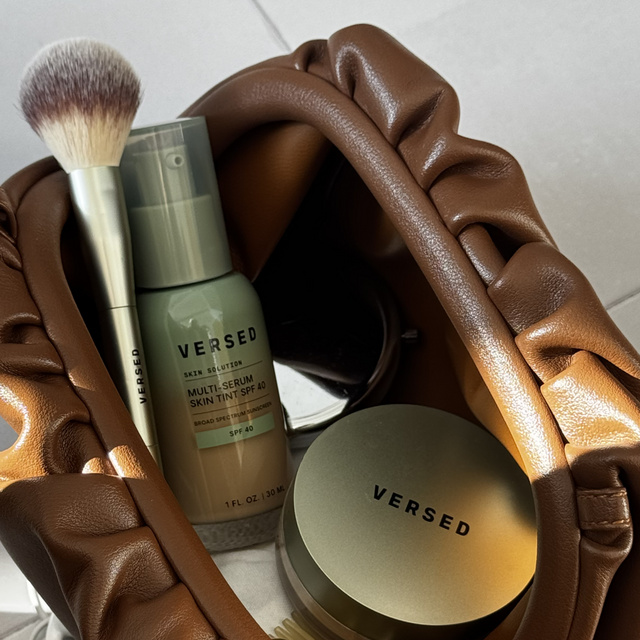 Brown sculptural hands holding a makeup brush, green tinted sunscreen bottle, and gold-toned compact product on white surface