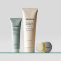 Three Versed skincare products: green microcrystal exfoliator tube, cream-colored AHA exfoliating body scrub tube, and small 